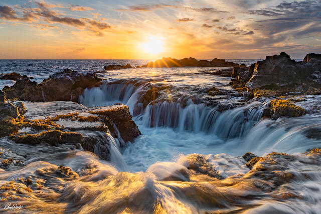 Ocean water flows through volcanic rock at sunset near Kona on the Big Island of Hawaii, with golden light reflecting across waves and a dramatic coast
