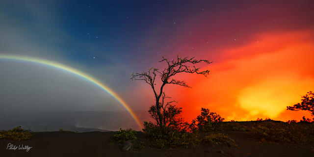 Moonbow arching across the night sky with glowing lava light and a silhouetted tree in Volcanoes National Park Hawaii.