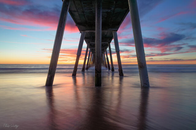 Sunset viewed from beneath the Manhattan Beach Pier with colorful clouds and smooth ocean water in California.