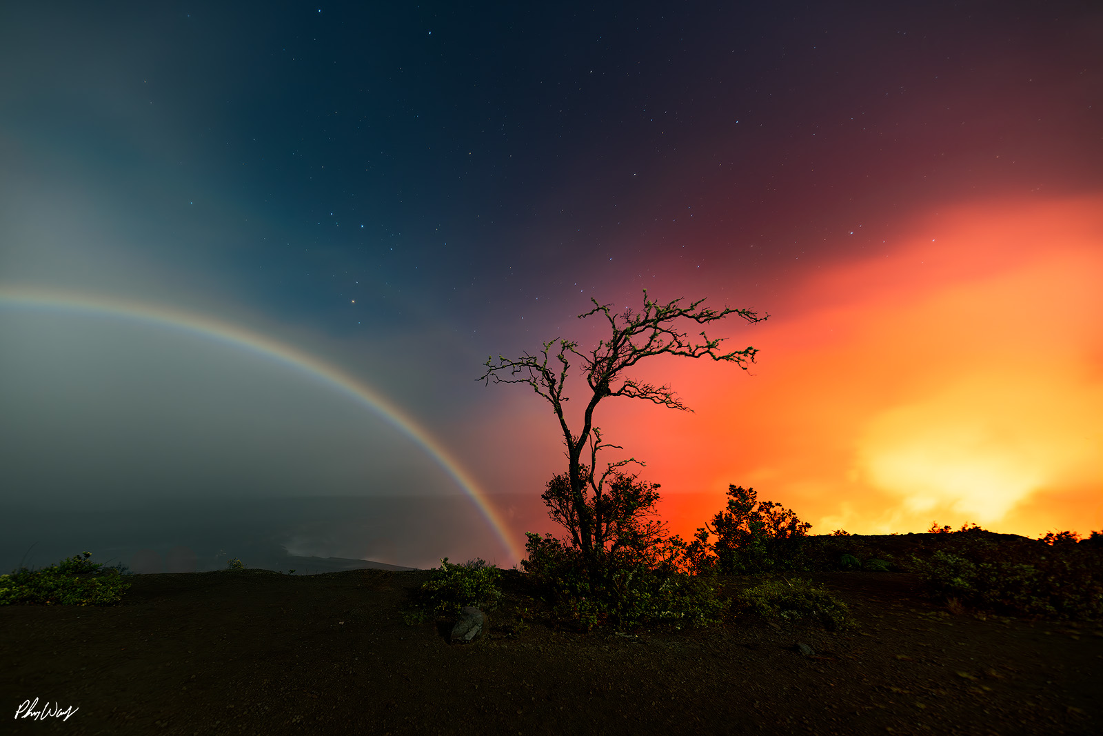Fire & Ice Moonbow arching across the night sky with glowing lava light and a silhouetted tree in Volcanoes National Park Hawaii.